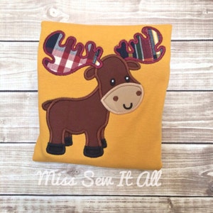 Op de afbeelding: Mosterdgeel kussen met een applicatie van een eland. De eland is bruin met een beige gezicht en zwarte hoeven. De geweien zijn geruit in rood, blauw en groen. De tekst "Miss Sew It All" staat onderaan.