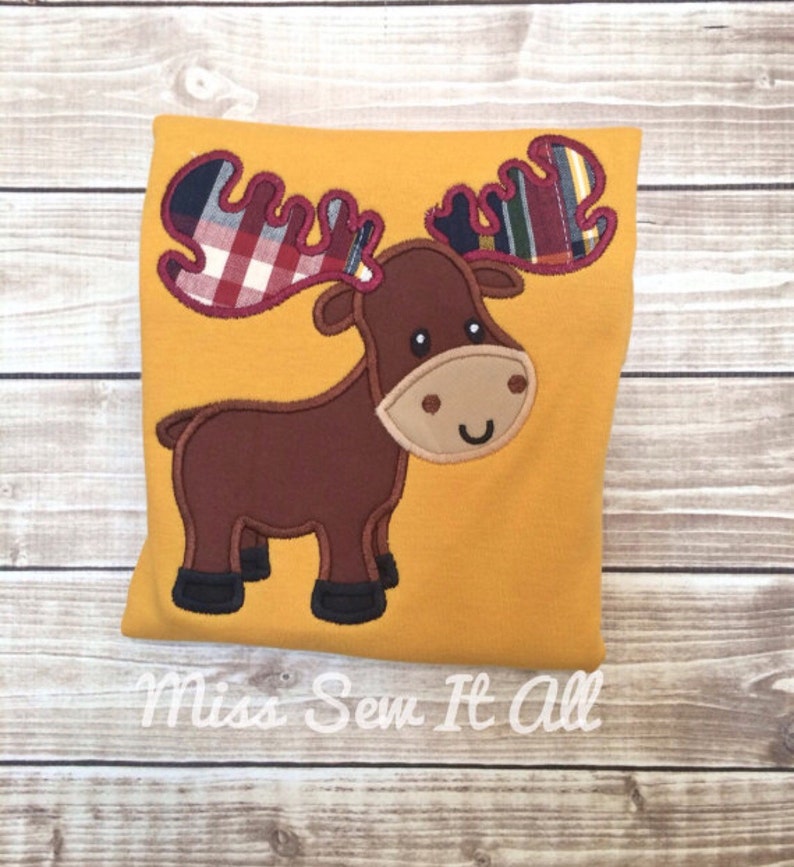 Op de afbeelding: Mosterdgeel kussen met een applicatie van een eland. De eland is bruin met een beige gezicht en zwarte hoeven. De geweien zijn geruit in rood, blauw en groen. De tekst "Miss Sew It All" staat onderaan.