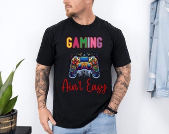 May include: Black t-shirt with a colorful graphic of a video game controller and the text "GAMING Ain't Easy" in rainbow and red colors. The design features vibrant details and a playful font, perfect for gamers.