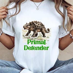 Op de afbeelding: Wit T-shirt met een groene en bruine illustratie van een ankylosaurus-dinosaurus. De tekst "Primal Defender" is in groen onder de dinosaurus gedrukt.