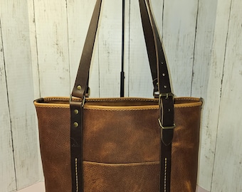 May include: A brown leather tote bag with a front pocket and brown leather straps. The bag has a distressed look and is made of high-quality leather.