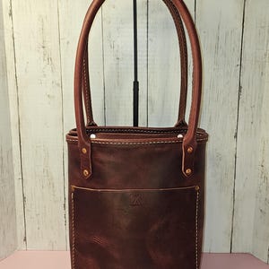 May include: A brown leather tote bag with a front pocket and a stitched design. The bag has brass rivets and a top handle.