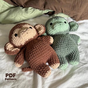 May include: Two crocheted monkey plushies, one brown and one green, lying on a white surface. The brown monkey has a beige face and the green monkey has a light green face. Both monkeys have black eyes and a stitched smile.