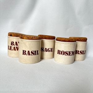 Può includere: Una collezione di sei barattoli di spezie in ceramica bianco sporco con coperchi in sughero. Ogni barattolo è etichettato con un nome di spezia diverso in lettere rosse: Bay Leaf, Basil, Sage, Rosemary e Parsley. I barattoli hanno un'estetica vintage e rustica.