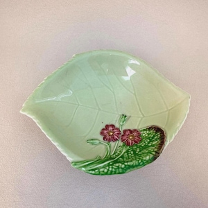 Può includere: Un piatto in ceramica verde chiaro a forma di foglia con venature a rilievo. Il piatto presenta un disegno in rilievo di due fiori rossi con steli e foglie verdi. Il piatto è probabilmente per uso decorativo.
