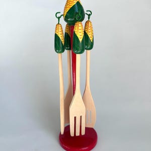 May include: A wooden kitchen utensil set with a red base and central support. The set includes a fork, two slotted spoons, and two solid spoons. The handles are topped with green and yellow corn-shaped designs.