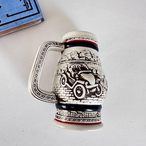 Op de afbeelding: Vintage keramische bierpul met een gedetailleerd reliëf van een klassieke auto en landschap. De pul heeft een witte basis met bruine, rode en blauwe accenten. De handgreep is versierd met een herhalend patroon. De tekst "1910 Stanley Steamer 1910" is zichtbaar.