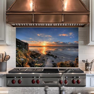 May include: A kitchen splashback depicting a sunset over a beach. The image shows a vibrant sky with orange and yellow hues reflecting on the water. The splashback is installed above a stainless steel gas range with red knobs.