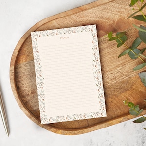 May include: A cream-coloured notepad with a floral border and the word "Notes" at the top, on a wooden tray. A silver pen is to the left. Eucalyptus sprigs are on the right. The notepad has lined pages.