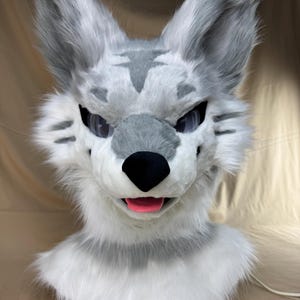 以下が含まれることがあります： グレーと白の毛皮のオオカミの頭。オオカミは黒い鼻、ピンクの舌、黒い目をしています。耳はグレーで尖っています。「Cosartcos」という文字が下にあります。