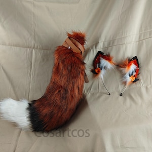 May include: A fluffy red and black fox tail with a white tip and a brown leather strap. A pair of fox ears with orange and white fur and black fabric accents.