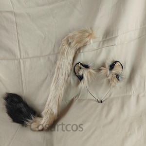 以下が含まれることがあります： フェイクファーの猫耳と尻尾のセット。耳は黒いヘッドバンドに付けられており、尻尾は長くふわふわとしたベージュと黒の尻尾です。