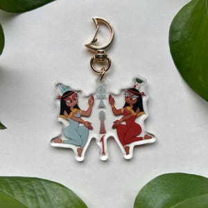 May include: A clear acrylic keychain featuring two stylized Egyptian figures in blue and red attire, with gold accents and a gold-colored clasp. The figures are holding their hands up, with an ankh symbol and hieroglyphs between them.