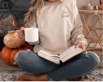 Puede incluir: Una sudadera beige con el texto "Over Stimulated" en letras negras. La sudadera la lleva puesta una persona sentada en el suelo con un libro y una taza.