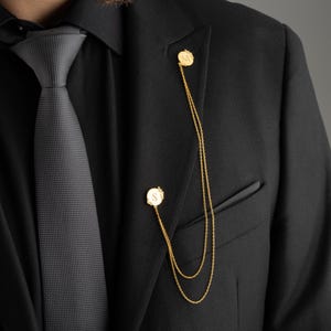 May include: A black suit jacket with a gray patterned tie and two gold chains with circular pendants. The pendants have an engraved letter 'S' on them.
