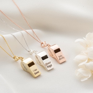 May include: Four personalized whistle necklaces in gold, silver, and rose gold. Each whistle pendant has a small rectangular opening and is engraved with a name and a phone number. The necklaces are displayed on a white, textured surface.