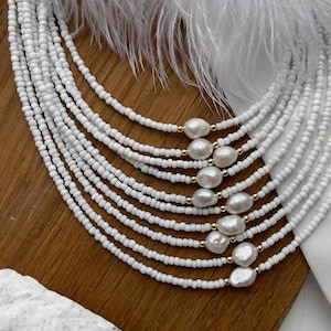 Op de afbeelding: Een witte kralenketting met meerdere strengen en grote witte parel accenten. De ketting wordt getoond op een houten oppervlak met een witte veer.