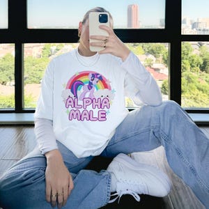 May include: White long-sleeved t-shirt featuring a pink and purple cartoon unicorn with a rainbow and clouds. The text "ALPHA MALE" is printed in pink letters below the unicorn.