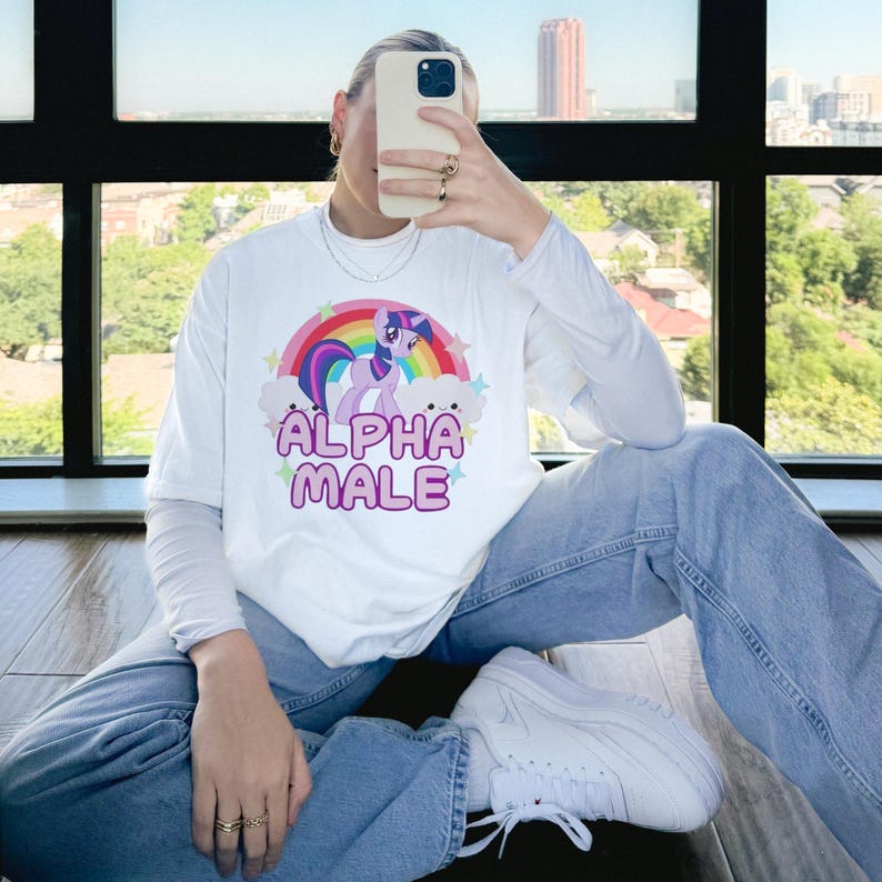 May include: White long-sleeved t-shirt featuring a pink and purple cartoon unicorn with a rainbow and clouds. The text "ALPHA MALE" is printed in pink letters below the unicorn.