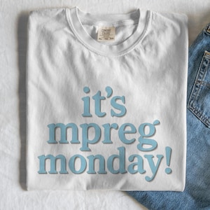 May include: White t-shirt with the text "it's mpreg monday!" in light blue. The shirt is folded and has a Comfort Colors tag. Part of a pair of blue jeans is visible in the corner.