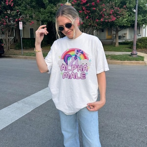 May include: White t-shirt featuring a graphic of a purple and pink cartoon unicorn with a rainbow and the text "ALPHA MALE" printed on it.