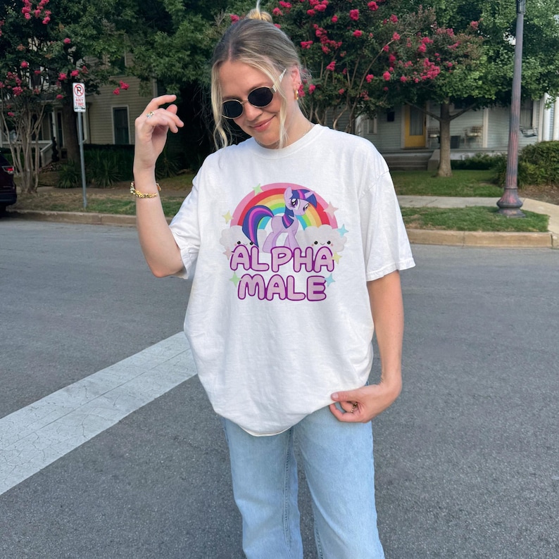 May include: White t-shirt featuring a graphic of a purple and pink cartoon unicorn with a rainbow and the text "ALPHA MALE" printed on it.