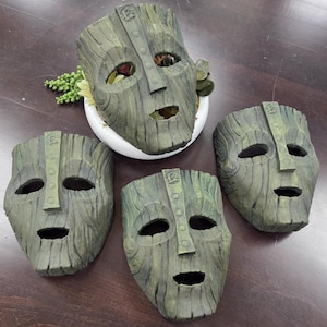 May include: Four wooden mask sculptures with a distressed, weathered finish. The masks have a carved appearance with eye and mouth openings. One mask is in a white planter with greenery. The masks are in shades of green and brown.