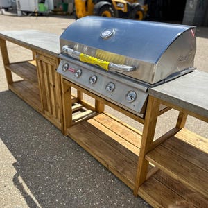 Może przedstawiać: Stalowy grill gazowy z czterema palnikami stoi na drewnianej wyspie kuchennej na zewnątrz z betonowym blatem. Wyspa ma po dwie p&oacute;łki z każdej strony.
