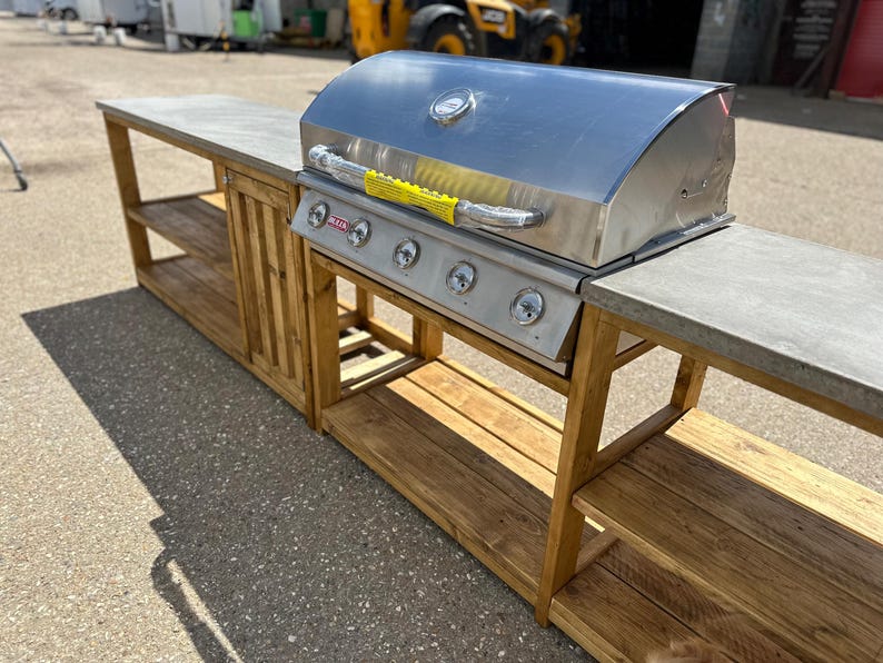 Może przedstawiać: Stalowy grill gazowy z czterema palnikami stoi na drewnianej wyspie kuchennej na zewnątrz z betonowym blatem. Wyspa ma po dwie p&oacute;łki z każdej strony.