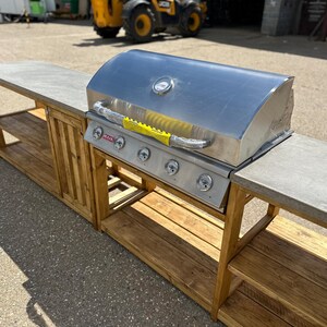 Może przedstawiać: Stalowy grill gazowy z pokrywą i czterema palnikami zamontowany na drewnianej ramie z betonowym blatem. Rama ma dwie p&oacute;łki po lewej stronie i dwie p&oacute;łki po prawej stronie.