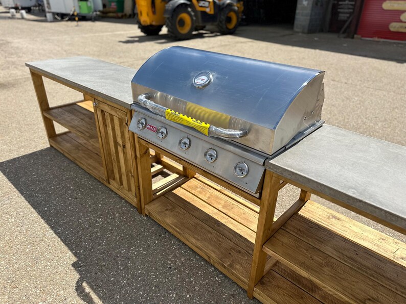 Może przedstawiać: Stalowy grill gazowy z pokrywą i czterema palnikami zamontowany na drewnianej ramie z betonowym blatem. Rama ma dwie p&oacute;łki po lewej stronie i dwie p&oacute;łki po prawej stronie.