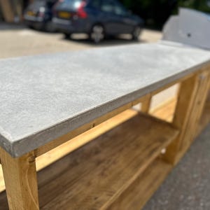 Może przedstawiać: Betonowy blat roboczy z gładkim, jasnoszarym wykończeniem spoczywa na drewnianym stole z dwoma p&oacute;łkami. St&oacute;ł wykonany jest z naturalnego drewna w jasnobrązowym kolorze.