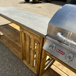 Może przedstawiać: Stalowy grill gazowy ze stalową obudową i drewnianym blatem. Grill posiada czerwone logo "BULL" i trzy pokrętła. Obudowa posiada drzwi z listwami i dwie p&oacute;łki.