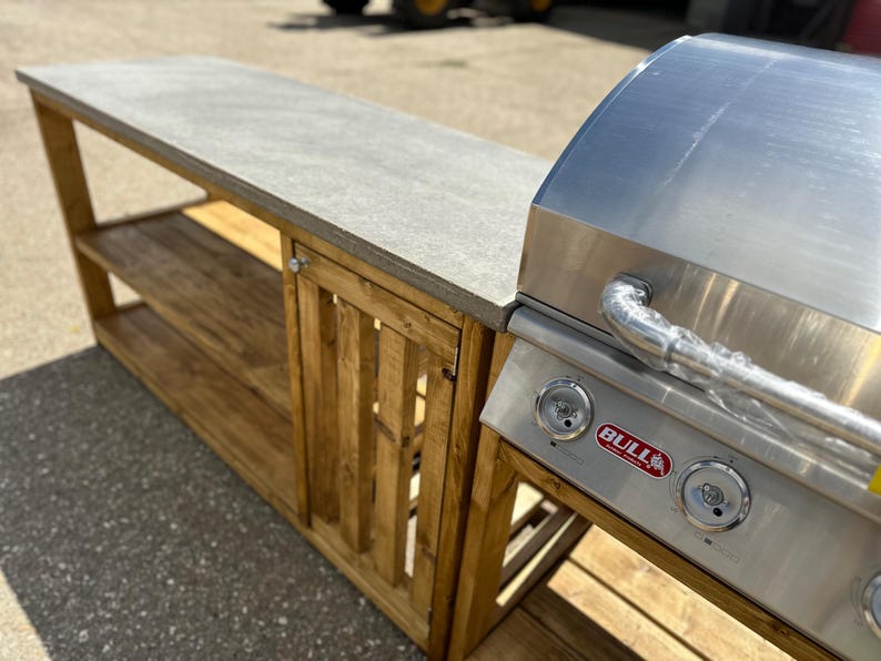 Może przedstawiać: Stalowy grill gazowy ze stalową obudową i drewnianym blatem. Grill posiada czerwone logo "BULL" i trzy pokrętła. Obudowa posiada drzwi z listwami i dwie p&oacute;łki.