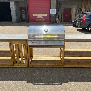 Może przedstawiać: Stalowy grill gazowy ze stali nierdzewnej z czterema palnikami i palnikiem bocznym, zamontowany na drewnianym blacie z dwoma p&oacute;łkami po obu stronach. Blat jest wykonany z betonu.