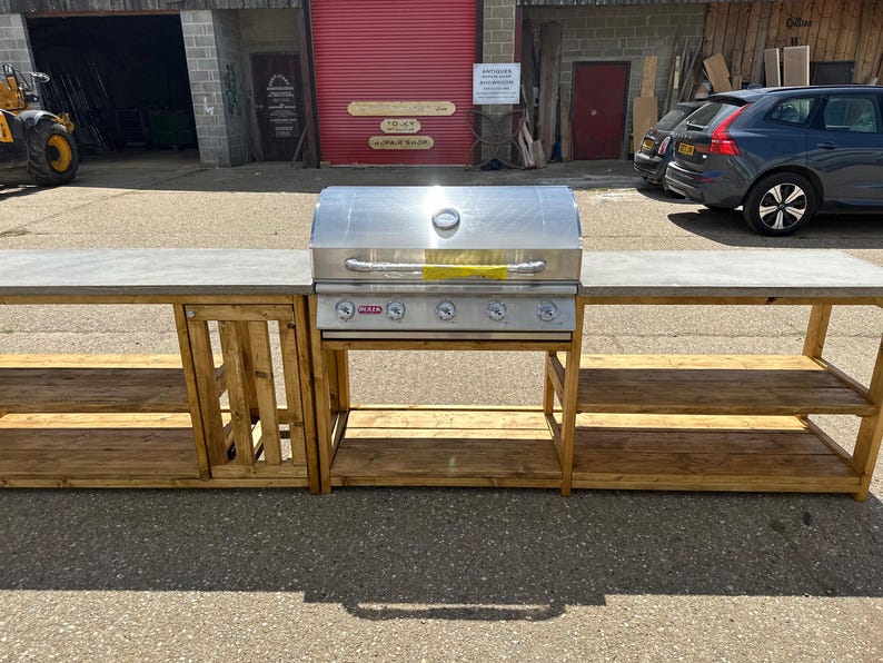 Może przedstawiać: Stalowy grill gazowy ze stali nierdzewnej z czterema palnikami i palnikiem bocznym, zamontowany na drewnianym blacie z dwoma p&oacute;łkami po obu stronach. Blat jest wykonany z betonu.