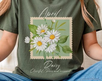 Op de afbeelding: Olijfgroen T-shirt met een bloemenontwerp van witte madeliefjes met gele centra en groene bladeren, geplaatst in een postzegelkader. De tekst luidt "April Daisy Cheerful, Pure and Innocent."