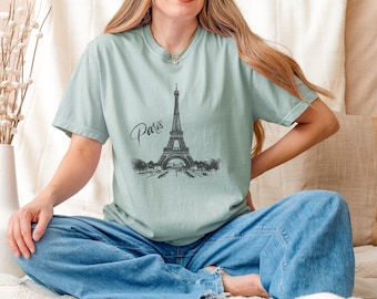 May include: A light blue t-shirt featuring a black and white illustration of the Eiffel Tower with the word "Paris" in cursive script. The shirt is made of a soft material and has short sleeves.