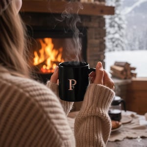 Può includere: Una tazza da caffè nera con la lettera "P" in un carattere serif, tenuta da una persona che indossa un maglione beige. Il vapore sale dalla tazza, con un camino e un paesaggio innevato sullo sfondo.