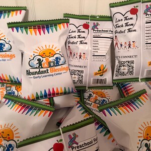 May include: A collection of white snack bags featuring the Abundant Blessings Early Learning Center logo with a sun, children, and crayons. The back displays Preschool Facts, a heart with an apple, and a QR code. The edges have green trim.