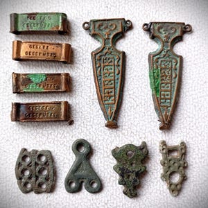 Könnte beinhalten: Eine Sammlung antiker Metallartefakte mit verwittertem Aussehen. Die Artikel umfassen rechteckige Stücke mit Text, schildförmige Anhänger und verschiedene andere dekorative Formen. Das Metall hat eine grün-braune Patina.