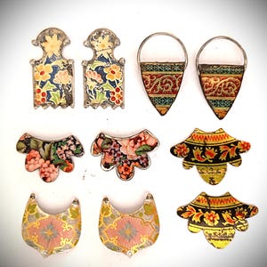 May include: A collection of ten decorative metal jewelry components featuring various shapes and colorful floral or geometric patterns. The set includes rectangular, triangular, and scalloped pieces with metallic edges and intricate printed designs.