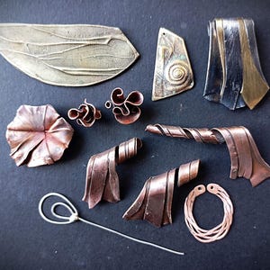 May include: A collection of metal jewelry components including a large brass wing-shaped piece, a spiral pendant, a two-tone metal pendant, a copper flower, two small copper swirls, two copper ribbon-like pieces, a silver wire pin, and a copper hoop.