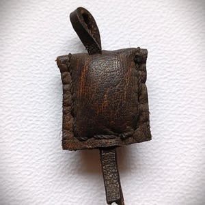 Könnte beinhalten: Ein kleiner, quadratischer Beutel aus dunkelbraunem Leder mit strukturierter Oberfläche. Der Beutel ist mit sichtbaren Nähten an den Kanten versehen und verfügt über eine Lederschlaufe zum Aufhängen sowie eine kurze Lederlasche an der Unterseite.