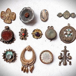 May include: A collection of antique jewelry pieces, including brooches and pendants, in various shapes and designs. The pieces feature gemstones, intricate metalwork, and a variety of colors, including gold, silver, red, and blue. The collection is displayed on a white surface.