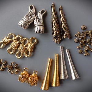 Könnte beinhalten: Eine Auswahl an Schmuckzubehör in Silber, Gold und Bronze. Die Abbildung zeigt Verschlüsse, konische Perlen, dekorative Kappen und andere Komponenten für die Schmuckherstellung. Die Teile sind auf einer grauen Oberfläche angeordnet.