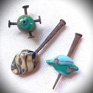 May include: Three handmade glass beads with nails. One bead is a green sphere with four nails, another is an oval with blue and cream swirls, and the third is a blue and green teardrop shape.
