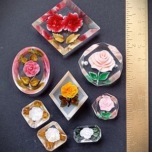 Könnte beinhalten: Eine Sammlung von Vintage-Blumen-Harz-Cabochons in verschiedenen Formen, darunter Diamanten, Ovale und Achtecke. Die Cabochons zeigen eingebettete Rosen und andere Blumen in Rosa, Rot, Gelb und Weiß. Ein Lineal befindet sich neben den Cabochons.