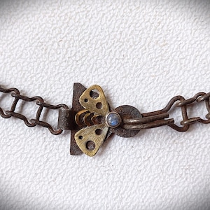 May include: Close-up of a necklace featuring a butterfly clasp. The clasp is made of brass and has a small, round, blue gemstone in the center. The chain is made of a dark, rusty metal. The butterfly clasp is the focal point of the necklace.