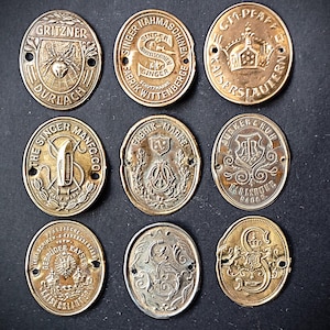 May include: Nine antique brass oval sewing machine badges with embossed text and designs. The badges feature various brand names, including Singer, Gritzner, and Pfaff, with intricate details and decorative elements.